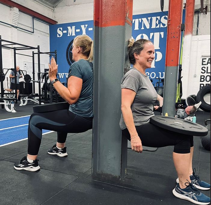 Two women doing a wall sit while holding weight plates during a full body HIIT class